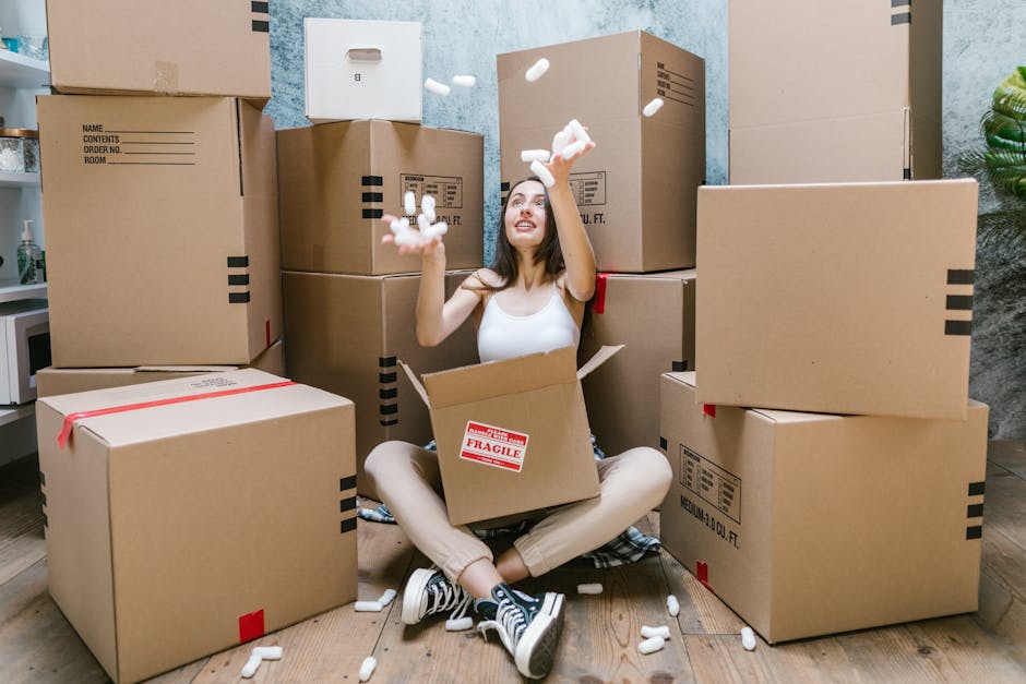 A young woman sitting cross-legged on a wooden floor surrounded by numerous large cardboard boxes, some labeled with fragile stickers, during a home relocation or furniture transport process. She is i